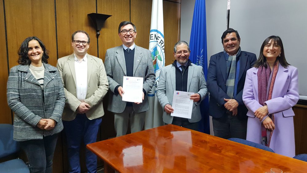 Asistentes a firma del convenio miran hacia el frente y las autoridades sostienen el convenio en sus manos.