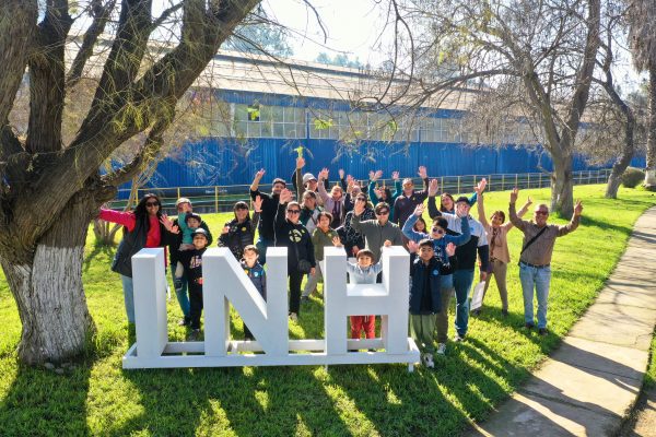 Grupo de personas de pie, saludando a la cámara con el laboratorio de fondo.